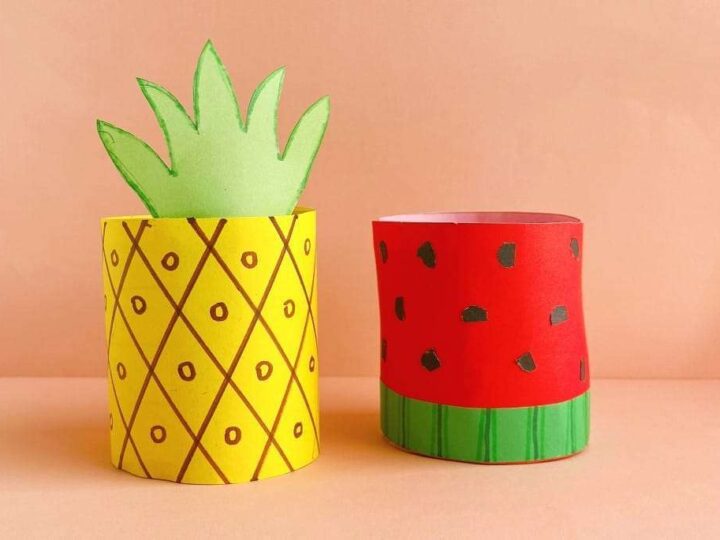 Two paper crafts on a peach-colored surface: one shaped like a pineapple with a yellow base and green leaves, and the other resembling a watermelon with a red base, black seeds, and a green stripe.
