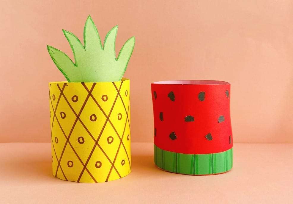 Two paper crafts on a peach-colored surface: one shaped like a pineapple with a yellow base and green leaves, and the other resembling a watermelon with a red base, black seeds, and a green stripe.