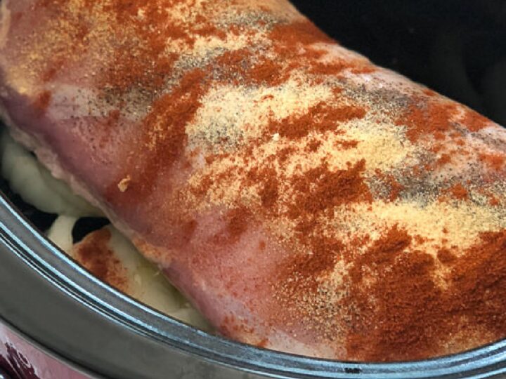 A seasoned pork loin sits atop sliced onions in a slow cooker. The meat is sprinkled with spices, including paprika and garlic powder, ready for cooking.