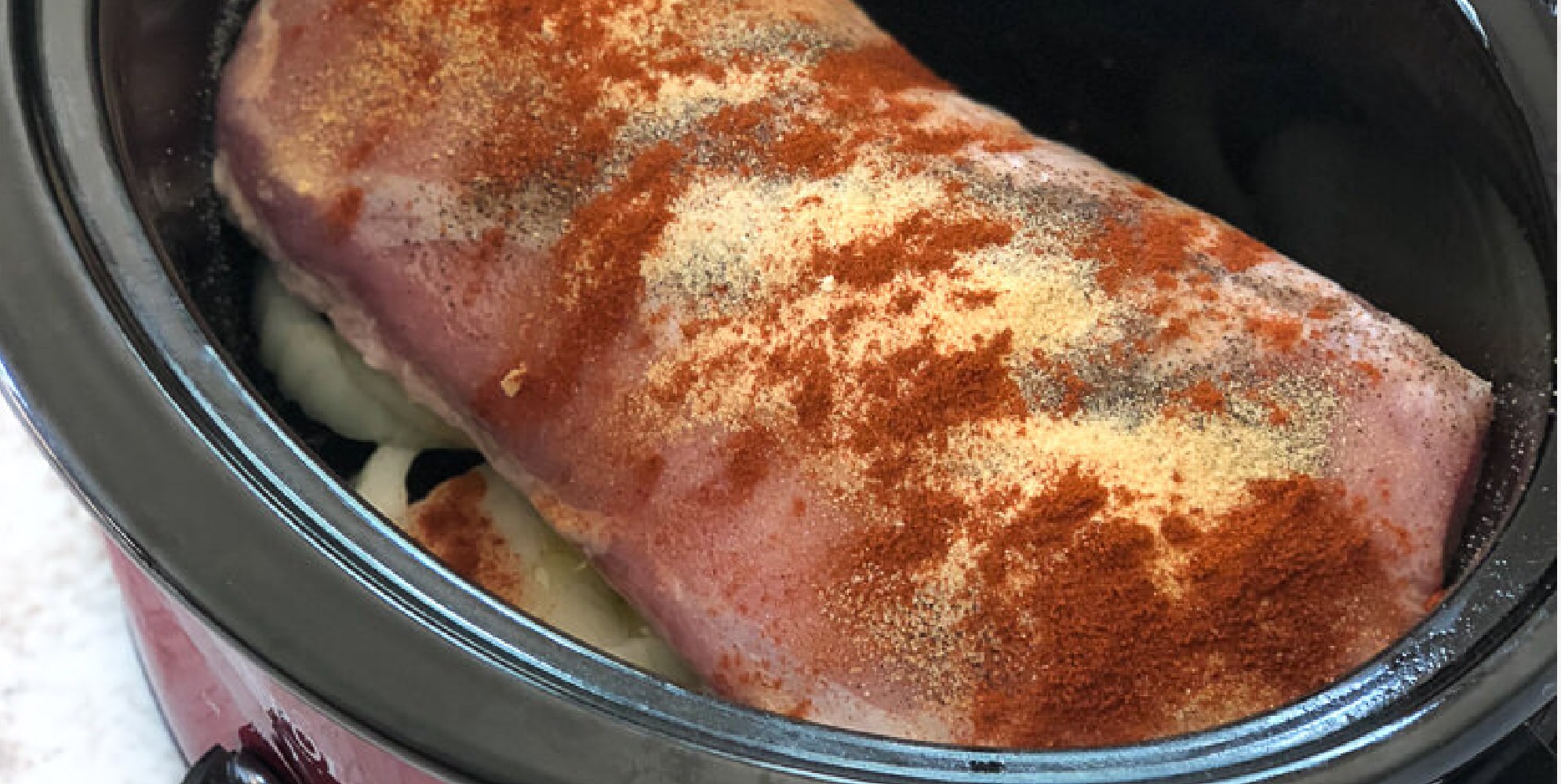 A seasoned pork loin sits atop sliced onions in a slow cooker. The meat is sprinkled with spices, including paprika and garlic powder, ready for cooking.