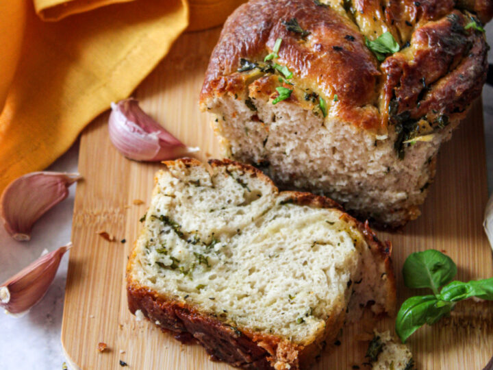 A loaf of herb and garlic bread on a wooden board, partially sliced, with fresh herbs and garlic cloves scattered around. A yellow cloth is partially visible in the background. The bread appears golden brown with green herbs throughout.