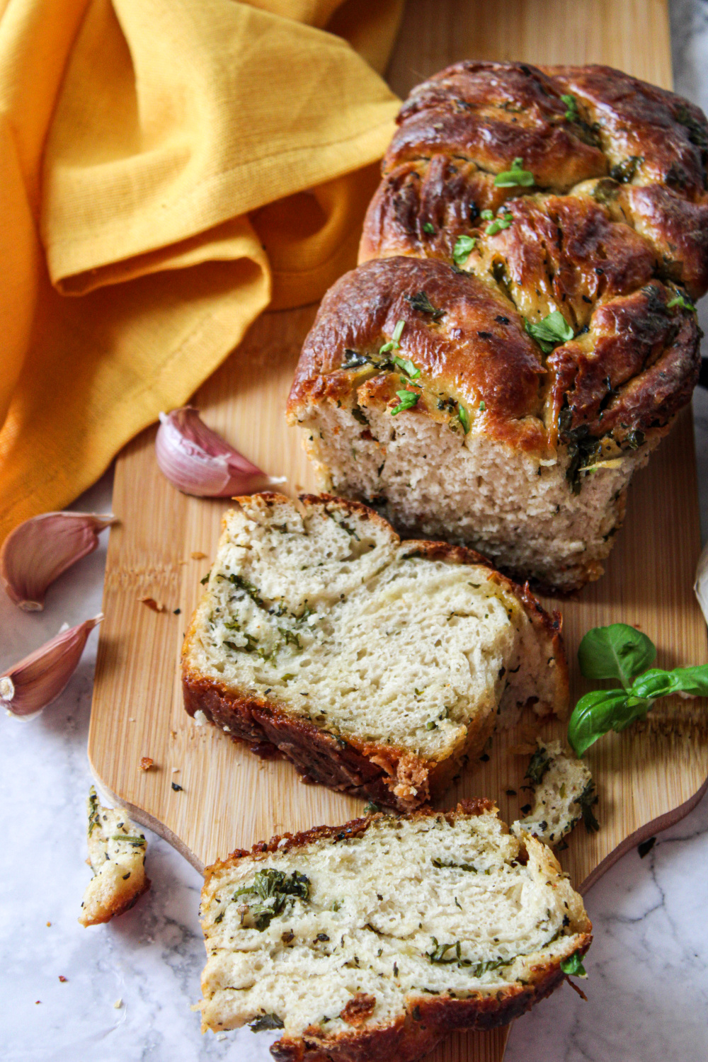 A loaf of herb and garlic bread on a wooden board, partially sliced, with fresh herbs and garlic cloves scattered around. A yellow cloth is partially visible in the background. The bread appears golden brown with green herbs throughout.