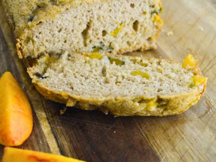 A loaf of peach bread on a wooden cutting board with one slice cut and placed in front. Two peach wedges are next to the slice. Fresh mint leaves are visible in the background.