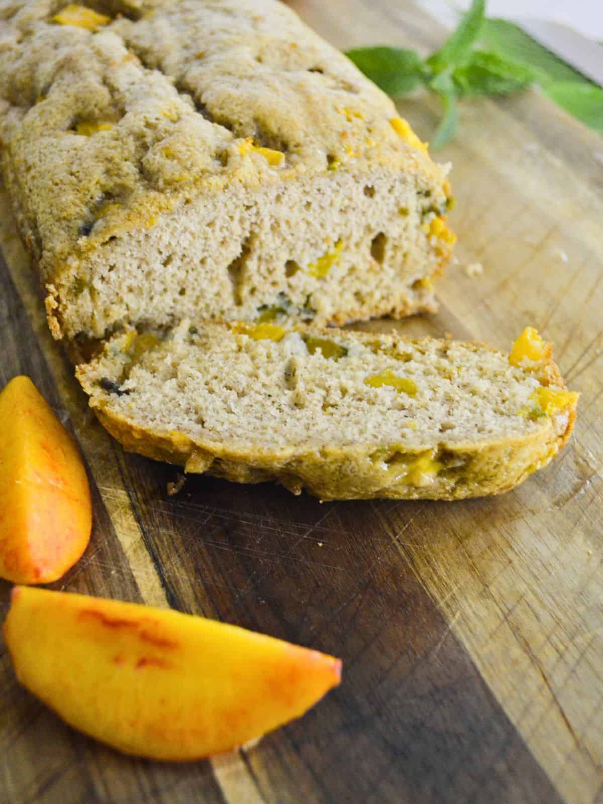 A loaf of peach bread on a wooden cutting board with one slice cut and placed in front. Two peach wedges are next to the slice. Fresh mint leaves are visible in the background.
