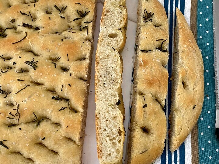 A freshly baked focaccia loaf on a white and blue striped napkin. The bread is sliced, showcasing a soft, airy interior with a golden crust topped with rosemary. A knife is partially visible next to the bread.