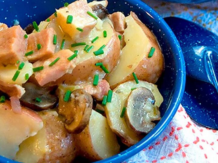 A blue bowl filled with a potato salad mixture, including chopped ham, sliced mushrooms, and diced potatoes, garnished with chopped chives. A blue spoon is placed beside the bowl on a colorful cloth.
