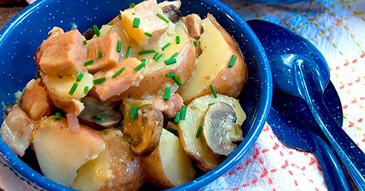 A blue bowl filled with a potato salad mixture, including chopped ham, sliced mushrooms, and diced potatoes, garnished with chopped chives. A blue spoon is placed beside the bowl on a colorful cloth.