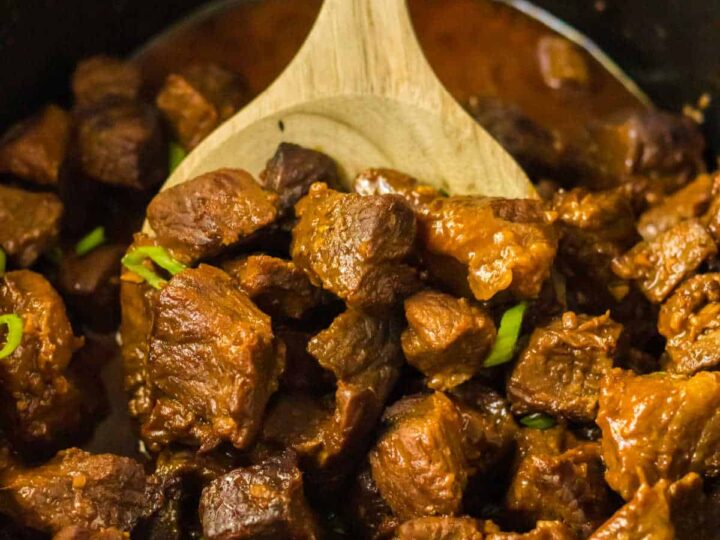 A wooden spoon scooping chunks of cooked beef stew from a pot. The beef is brown and tender, garnished with small pieces of green herbs. The stew appears rich and flavorful, with a thick, dark sauce.