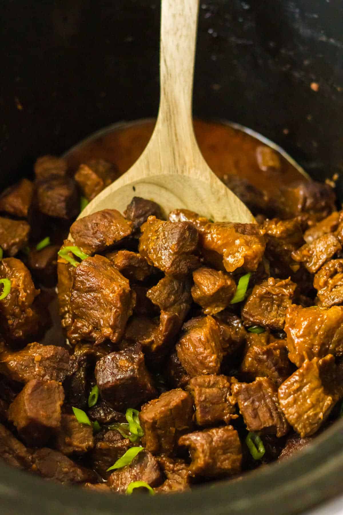 A wooden spoon scooping chunks of cooked beef stew from a pot. The beef is brown and tender, garnished with small pieces of green herbs. The stew appears rich and flavorful, with a thick, dark sauce.