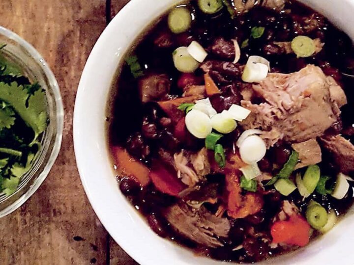 A bowl of chili with chunks of meat, black beans, red bell peppers, and topped with chopped green onions. Beside the bowl is a small dish of fresh cilantro. Both are on a wooden table.