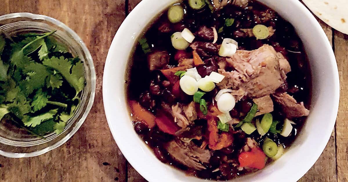 A bowl of chili with chunks of meat, black beans, red bell peppers, and topped with chopped green onions. Beside the bowl is a small dish of fresh cilantro. Both are on a wooden table.