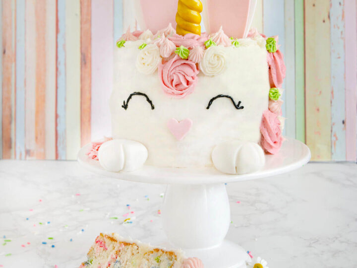 A colorful unicorn-themed cake with a golden horn, pink ears, and floral decorations sits on a white cake stand. A slice of the cake, showing sprinkle-filled layers, is on a plate below. The background is a pastel-striped wall.