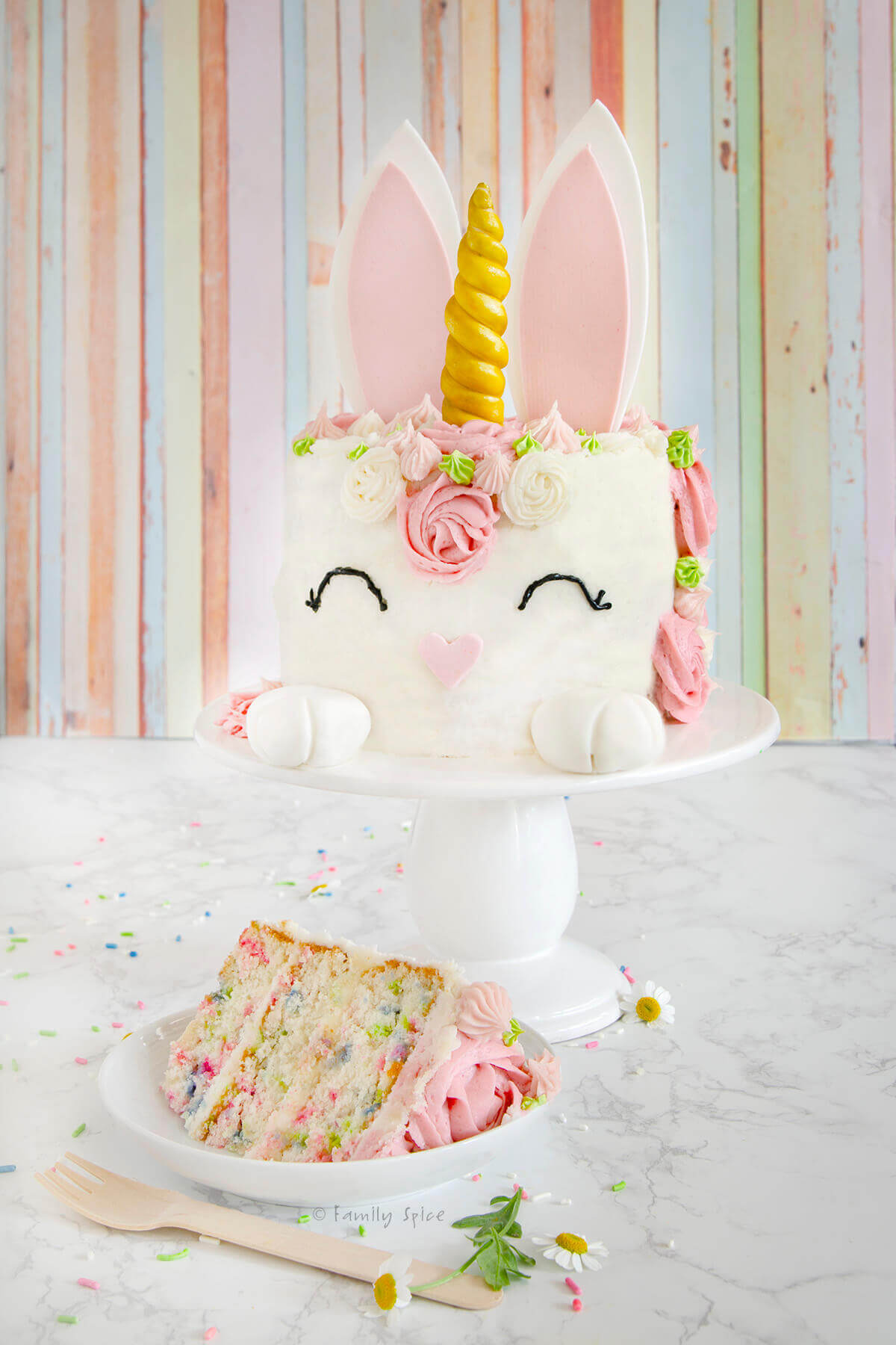A colorful unicorn-themed cake with a golden horn, pink ears, and floral decorations sits on a white cake stand. A slice of the cake, showing sprinkle-filled layers, is on a plate below. The background is a pastel-striped wall.