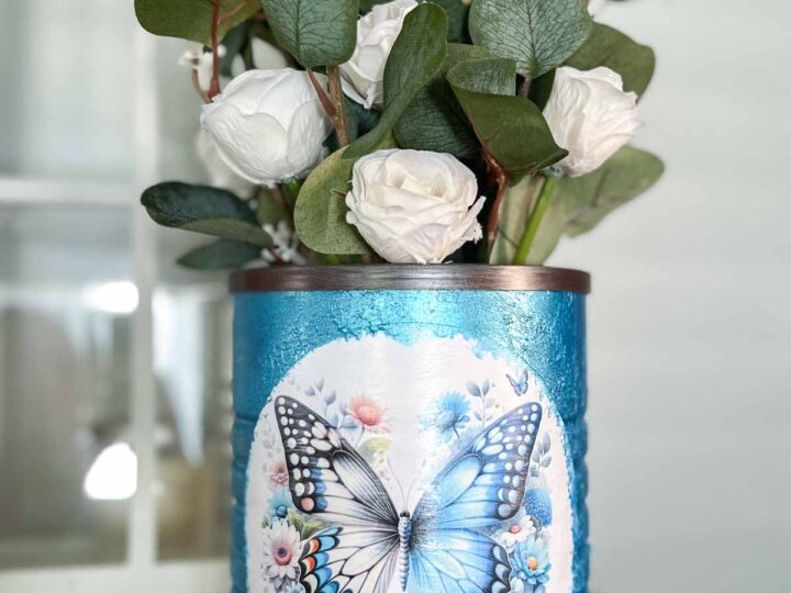 A blue and white decorative vase contains white roses and green leaves. The vase features a large butterfly and floral design on the front. It is placed on a wooden surface with a window in the background.