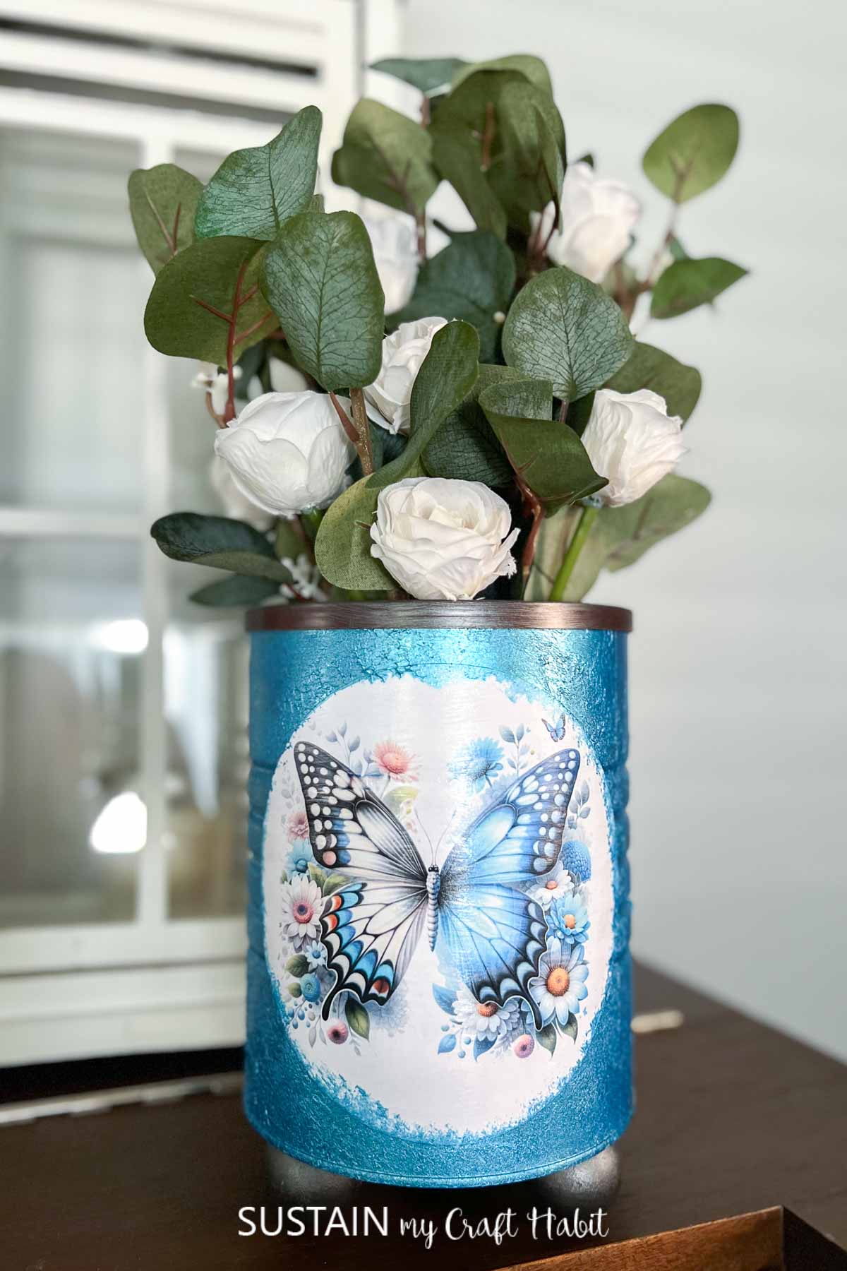 A blue and white decorative vase contains white roses and green leaves. The vase features a large butterfly and floral design on the front. It is placed on a wooden surface with a window in the background.