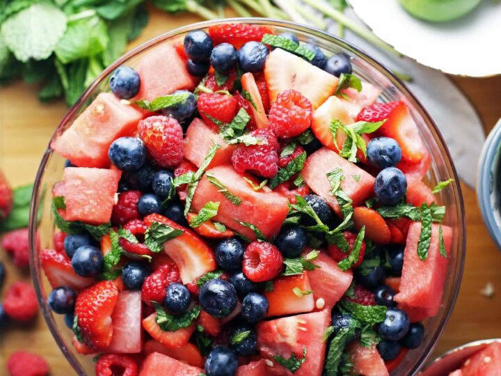 A bowl of mixed fresh fruit including watermelon cubes, strawberries, blueberries, and raspberries, garnished with mint leaves. Surrounding the bowl are additional fruits, mint sprigs, a lime, and a knife on a wooden surface.