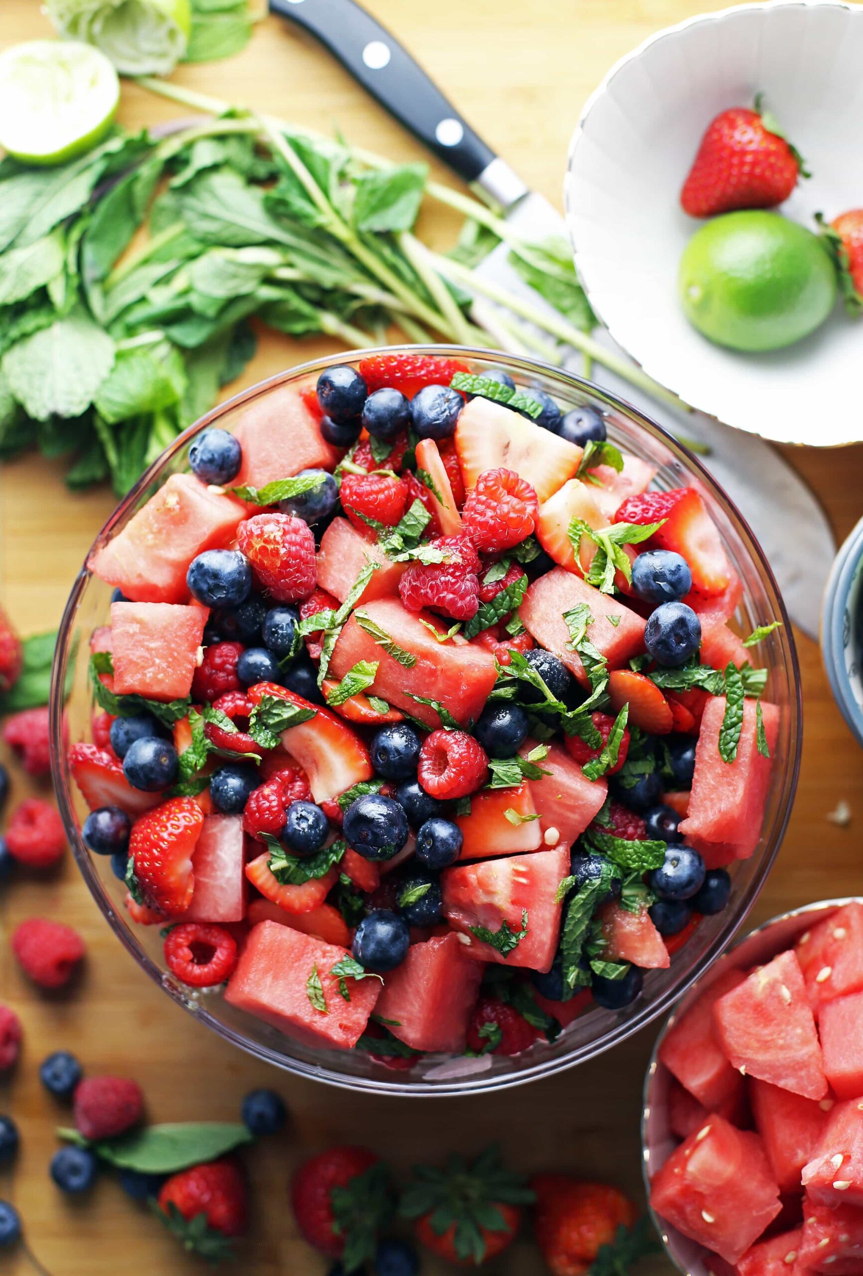 A bowl of mixed fresh fruit including watermelon cubes, strawberries, blueberries, and raspberries, garnished with mint leaves. Surrounding the bowl are additional fruits, mint sprigs, a lime, and a knife on a wooden surface.