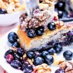 A slice of blueberry walnut coffee cake, inspired by our Blueberry Walnut Coffee Cake Recipe, sits on a white plate with a fork inserted. Fresh blueberries and walnuts are scattered around, adding extra flavor to each bite.