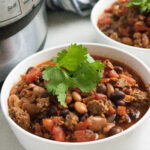 Two bowls of chili, filled with beans, ground meat, tomatoes, and garnished with cilantro, are placed beside an electric pressure cooker. A striped cloth is in the background.