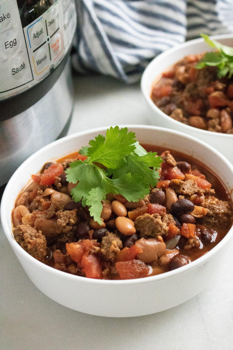 Two bowls of chili, filled with beans, ground meat, tomatoes, and garnished with cilantro, are placed beside an electric pressure cooker. A striped cloth is in the background.