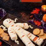 Mummy cheese ball, fun recipe: a cheese mummy with piped cream cheese bandages on a tray with crackers and fall leaves
