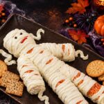 Cheese mummy with piped cream cheese bandages, peppercorn eyes and a pimiento mouth on a dark tray with crackers and fall leaves