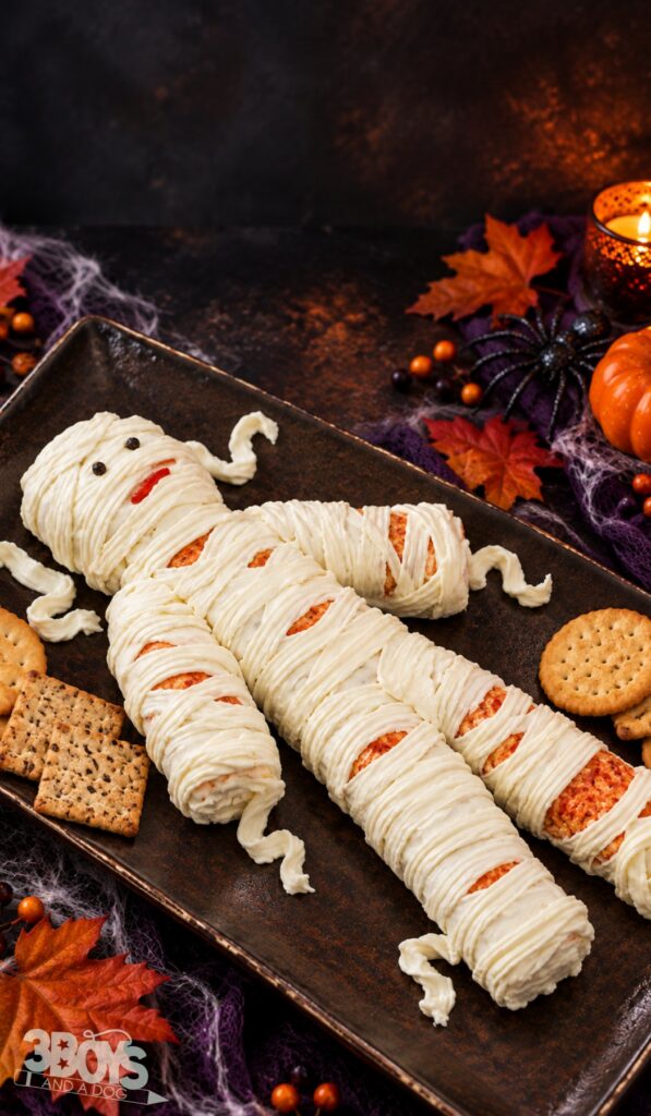 Cheese mummy with piped cream cheese bandages, peppercorn eyes and a pimiento mouth on a dark tray with crackers and fall leaves