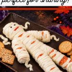 Mummy Cheese Ball, perfect for sharing: a cheese mummy with cream cheese bandages on a dark tray surrounded by crackers and autumn leaves