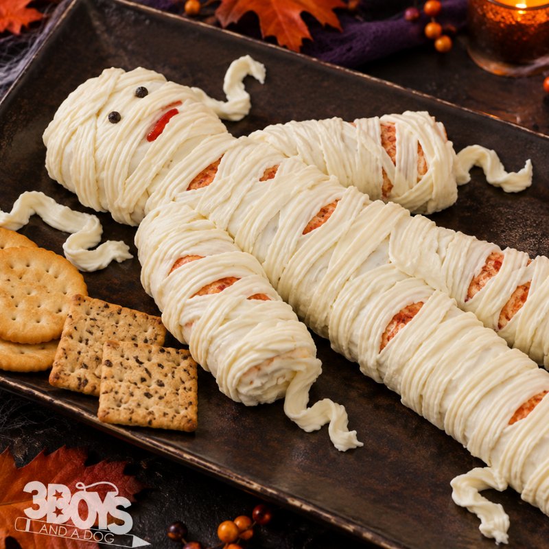 Close-up of a cheese mummy with piped cream cheese bandages over port wine cheese logs, served with crackers
