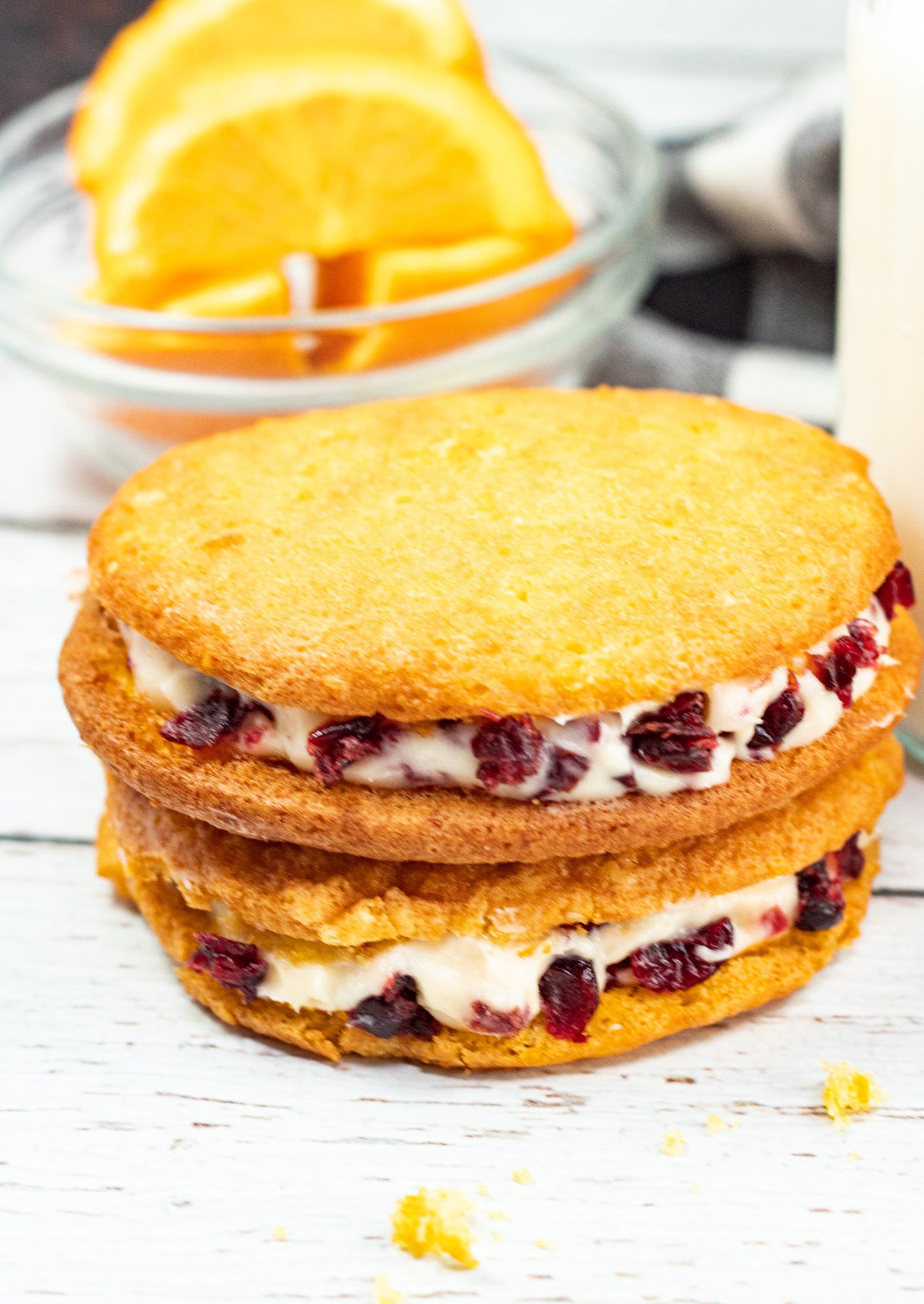 Three cranberry orange whoopie pies stacked on a white plate, cream cheese filling studded with dried cranberries, orange slices behind