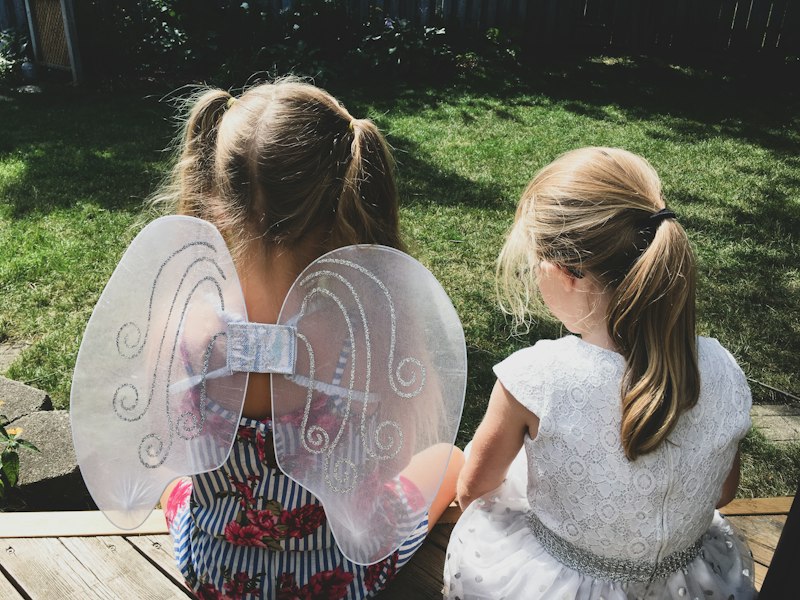 Two little girls sitting outside, one wearing white fairy wings