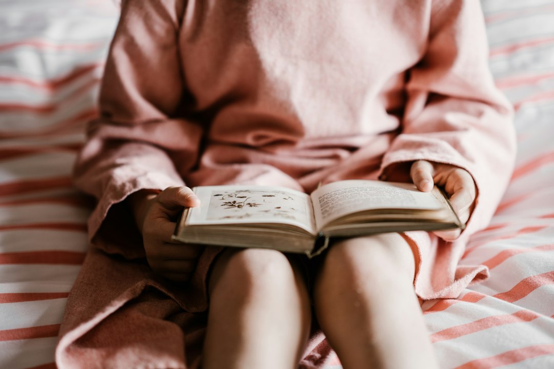 Little girl in pink pajamas reading a storybook on her bed